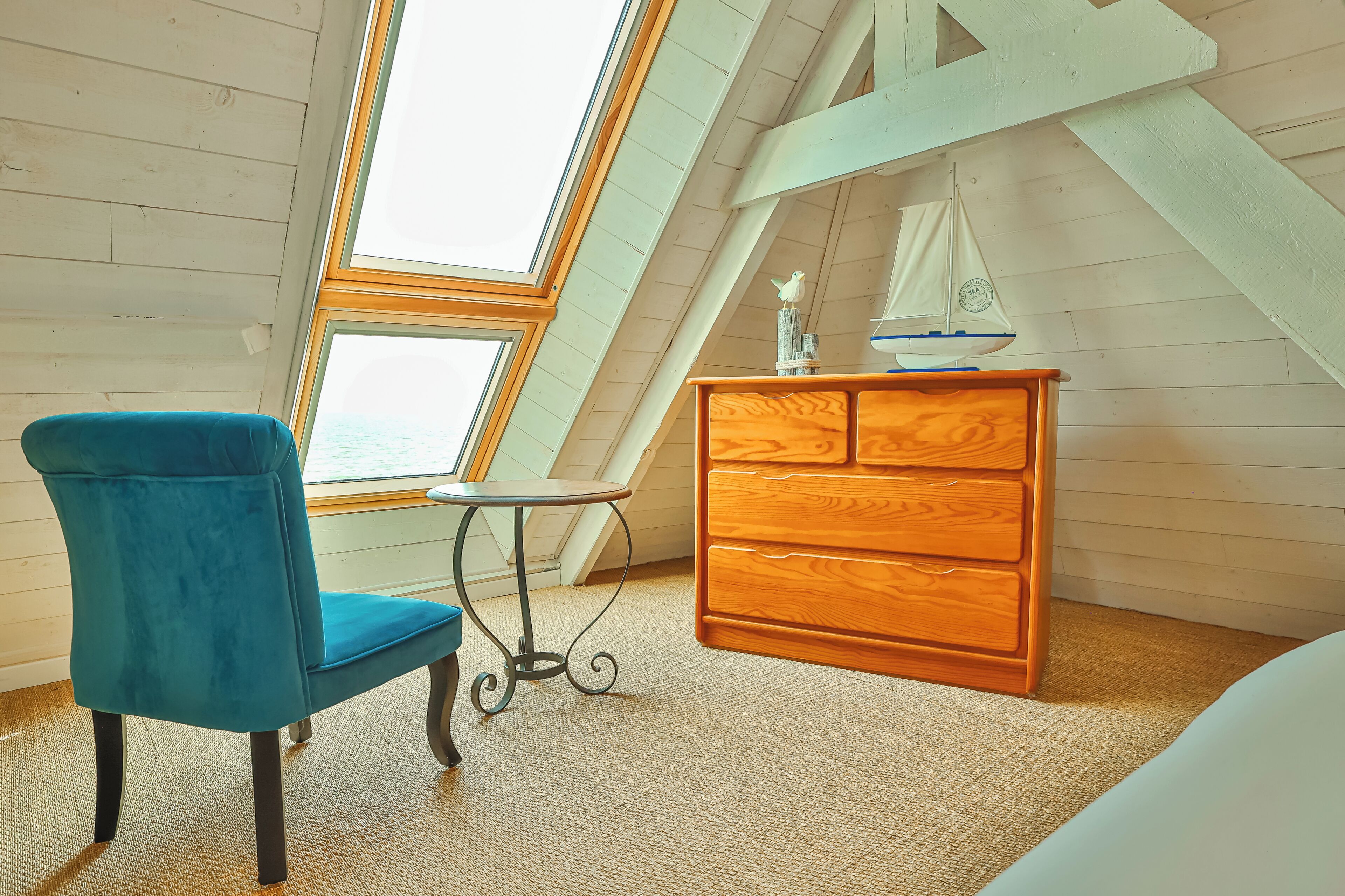 Een kamer met een schuine kap, een houten dressoir, een blauwe stoel en een kleine tafel.