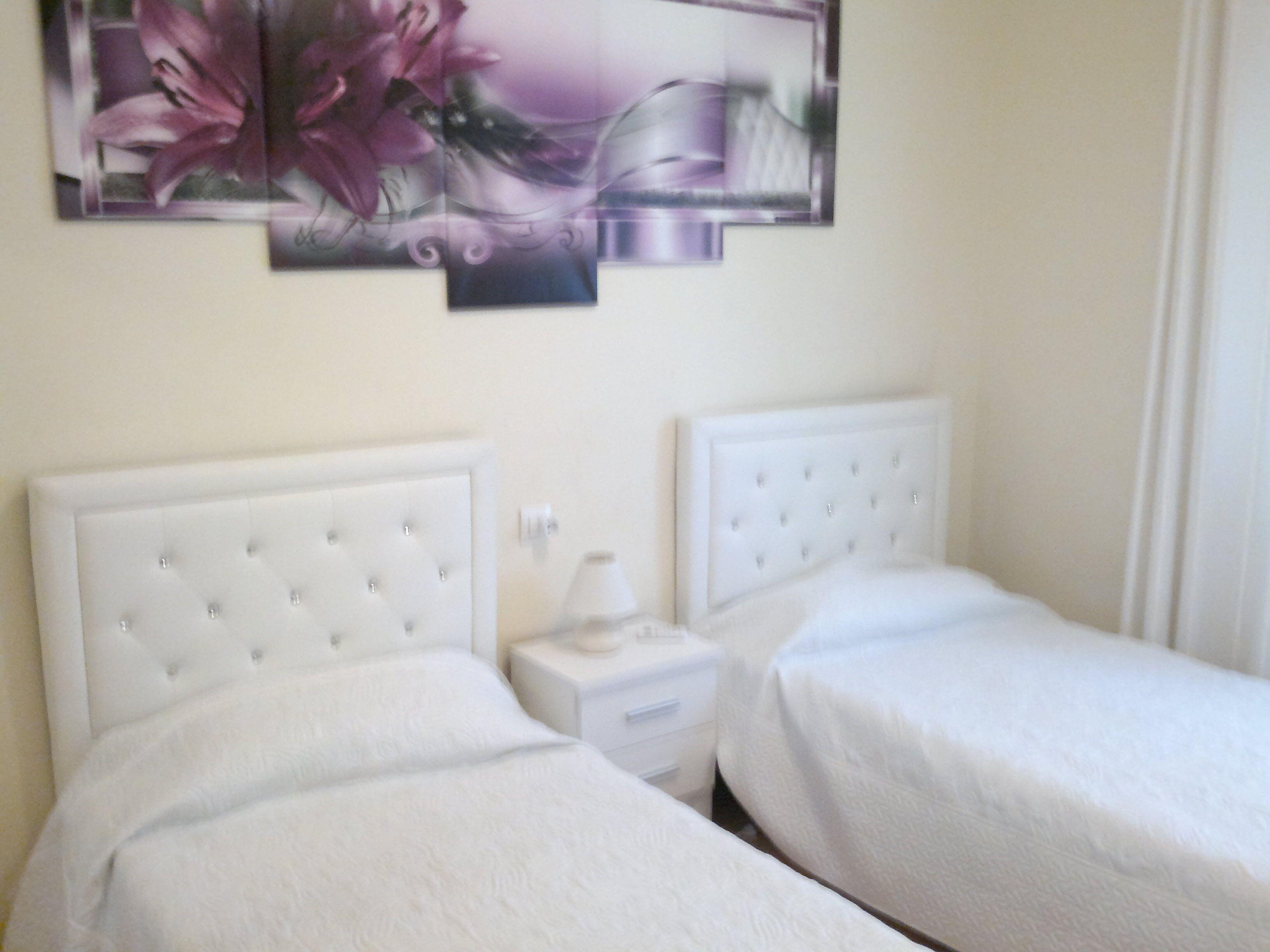 Una habitación de hotel con dos camas, cada una con cabecero y cubrecama blancos. Hay una mesita de noche entre las camas con una lámpara y una pequeña mesa.