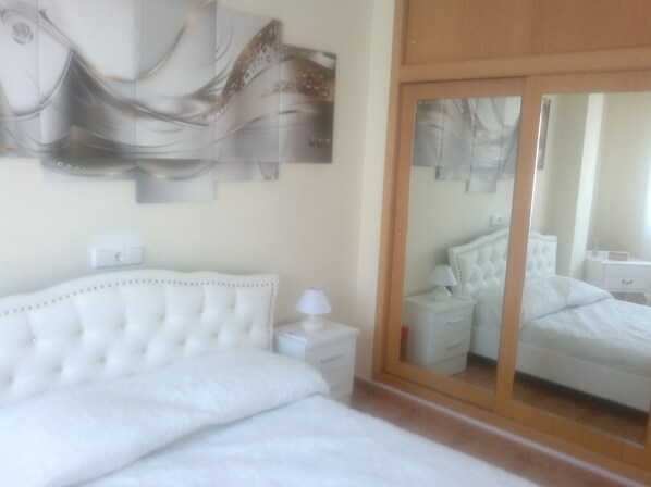 A bedroom with a white bed, a nightstand, a lamp, and a wall-mounted abstract art piece.