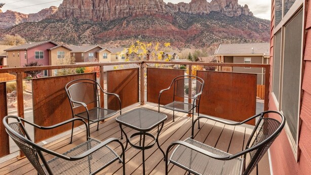 Ein Balkon mit Metallstühlen und Tischen, mit Blick auf eine gebirgige Landschaft und Häuser im Hintergrund.