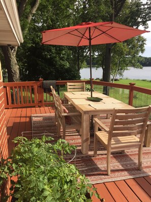 Eine Holzterrasse mit Tisch und Stühlen, einem roten Sonnenschirm und Blick auf einen See.