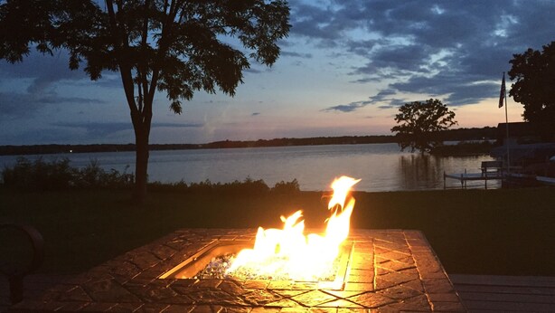 Un hogar con fuego, rodeado de una zona pavimentada con vista a un lago y árboles al anochecer.