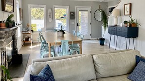 En moderne stue med en beige sofa, et spiseområde med et træbord og blå stole, en pejs og et køkken, der kan ses igennem en åben dør.