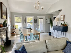 A modern living room with a beige sofa, a dining area with a wooden table and blue chairs, a fireplace, and a kitchen visible through an open door.