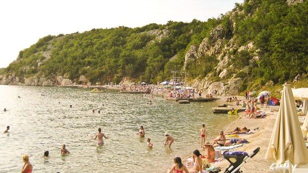 A beach with people swimming and sunbathing, surrounded by greenery and rocky cliffs.