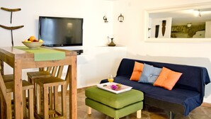 A living room with a wooden dining table, a green ottoman, a blue sofa with orange cushions, a flat-screen TV, and a small vase on the wall.