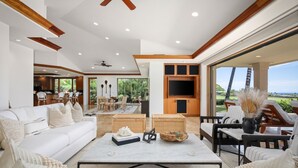 A spacious living area with a white sofa, coffee table, and a dining area with chairs. There is a TV mounted on the wall and a view of the outdoors.