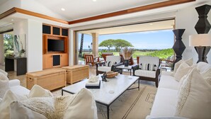 A spacious living area with a large window offering a view of a lush landscape. There is a seating area with a sofa and chairs, a coffee table, and a television mounted on the wall.