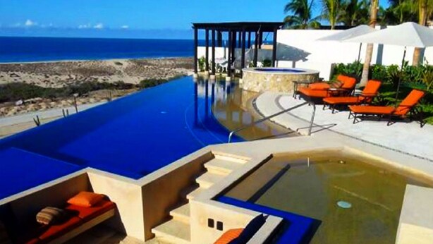 Zona de piscina con vista despejada al océano, un espacio de descanso cubierto y sillas de lounge color naranja.