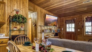 Un intérieur en bois avec une table à manger dressée pour deux personnes, une bibliothèque et un téléviseur diffusant un paysage pittoresque.
