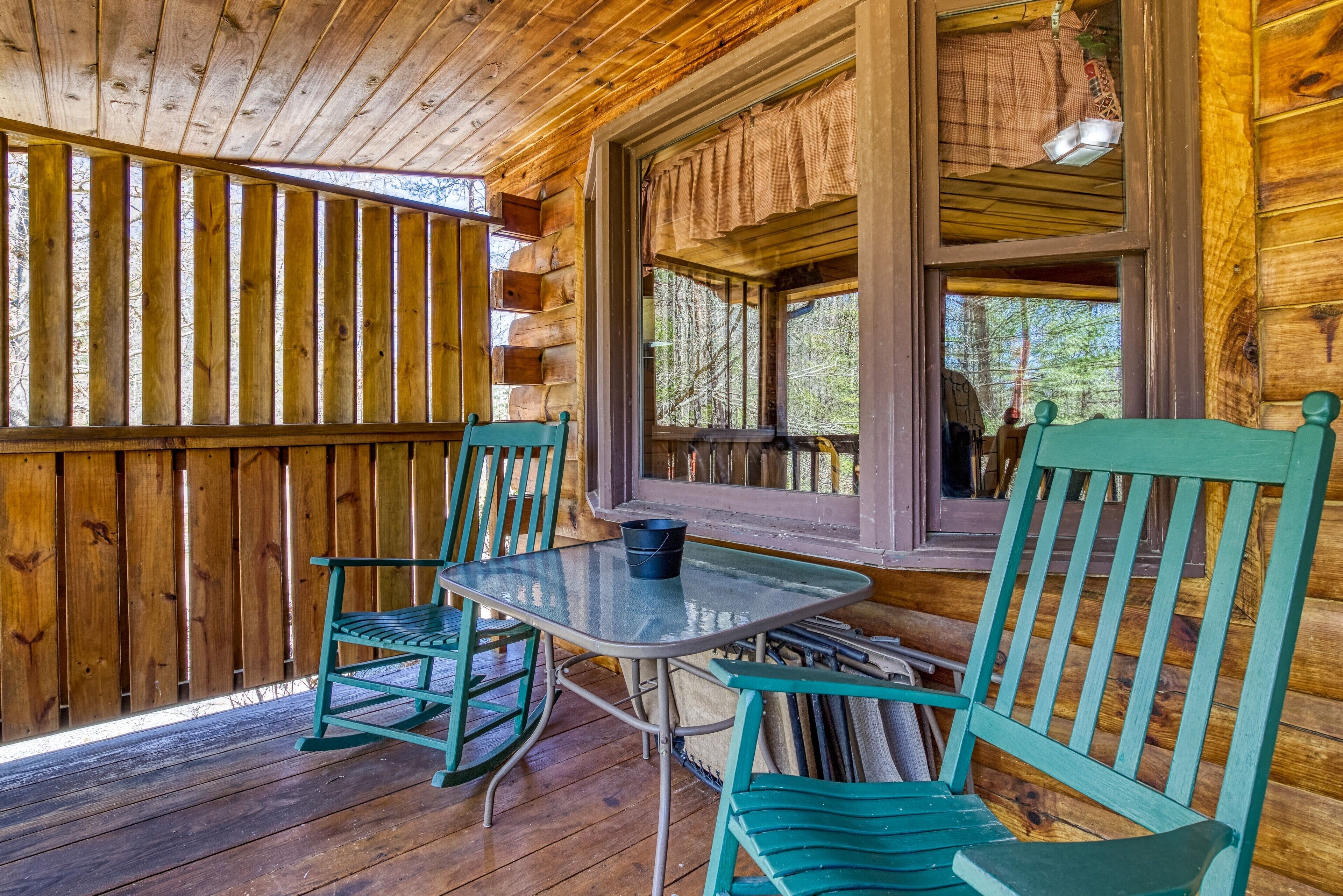 A wooden cabin with a covered porch, two green chairs, a table, and a window with curtains.