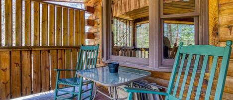 A wooden cabin with a covered porch, two green chairs, a table, and a window with curtains.