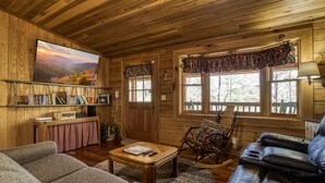 L’intérieur chaleureux d’un chalet, avec un revêtement mural en bois, un téléviseur fixé au mur, une bibliothèque, un fauteuil à bascule et un canapé en cuir.
