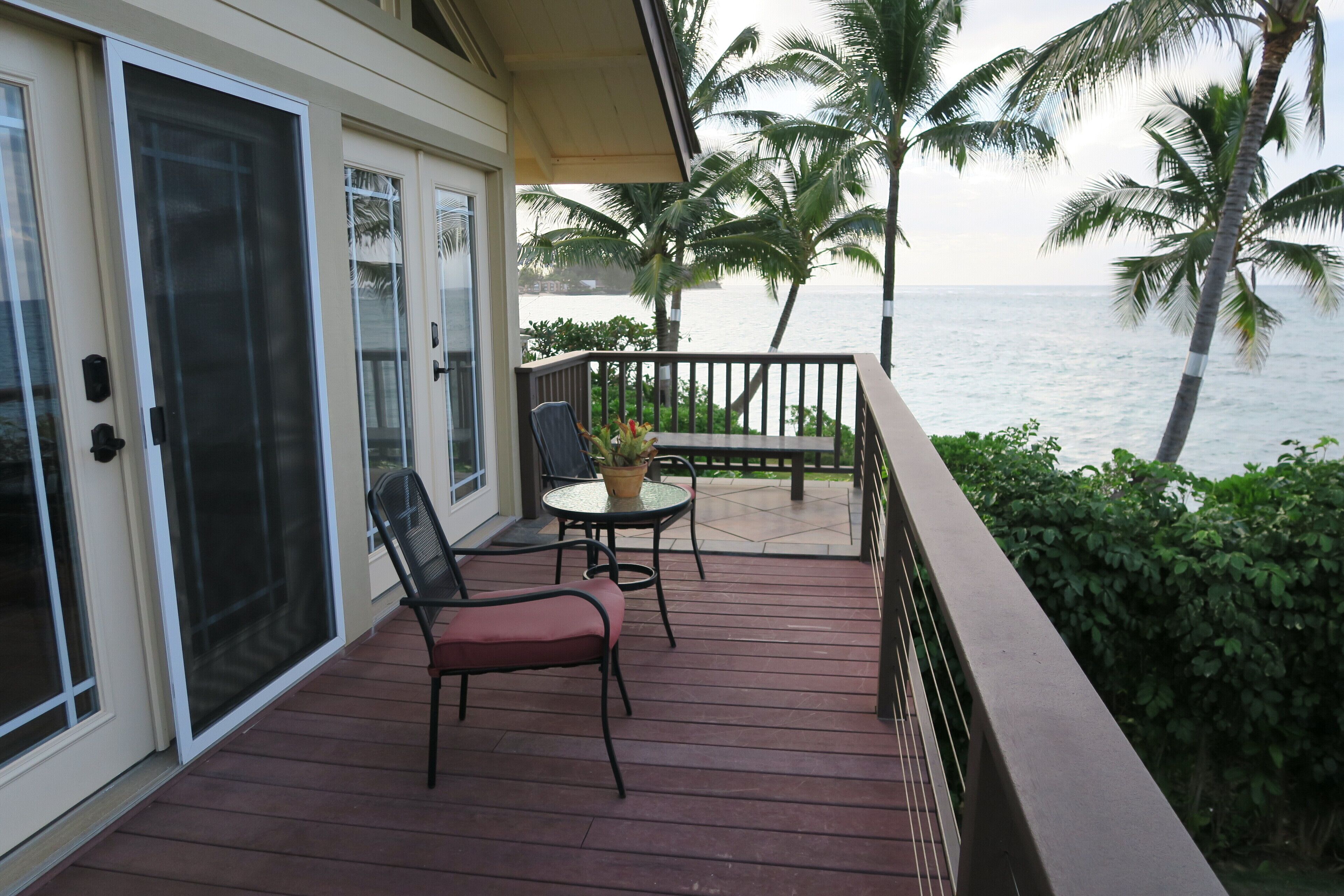 A balcony with a red wooden floor, two chairs, a small table with a plant, and a view of the ocean with palm trees.