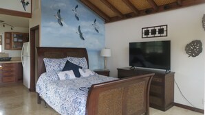 A bedroom with a bed, a TV, a wall with bird art, and a kitchen in the background.