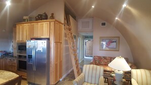 A compact kitchen with wooden cabinets, a built-in oven, and a refrigerator. A ladder is propped against the wall, and a sofa with a floral pattern is in the adjacent living area. - Conch House on a Quiet Canal : Monthly Only (Little Torch Key)