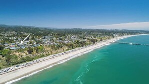 Vista aérea de un pueblo costero con playa, muelle y una carretera bordeada de coches aparcados.