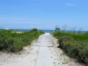 A concrete pathway leading to the beach, flanked by grassy areas and a clear blue sky.