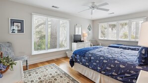A bedroom with two beds, a ceiling fan, a TV, and large windows with blinds.