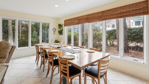 A dining room with a large table set for six, surrounded by wooden chairs with black cushions. The room has large windows with a view of greenery and buildings outside.