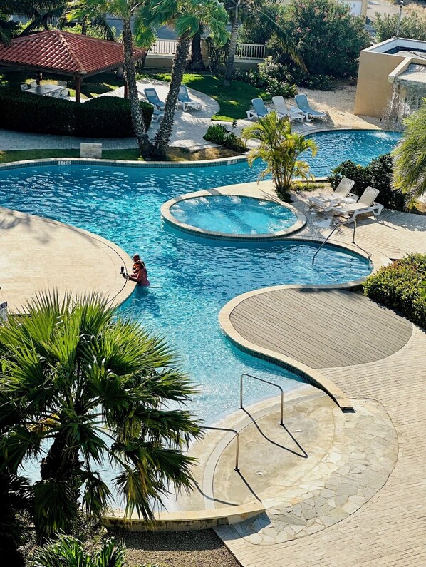 A pool area with multiple water features, including a hot tub and a circular pool, surrounded by lounge chairs and palm trees. - COZY EAGLE BEACH VIEW CONDO (Oranjestad west)
