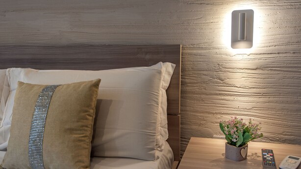 A neatly made bed with a beige pillow and a decorative pillow with a silver trim. A small potted plant with pink flowers is on a bedside table next to a remote control. - Casa Vacanze Carolina 348 (Naples)