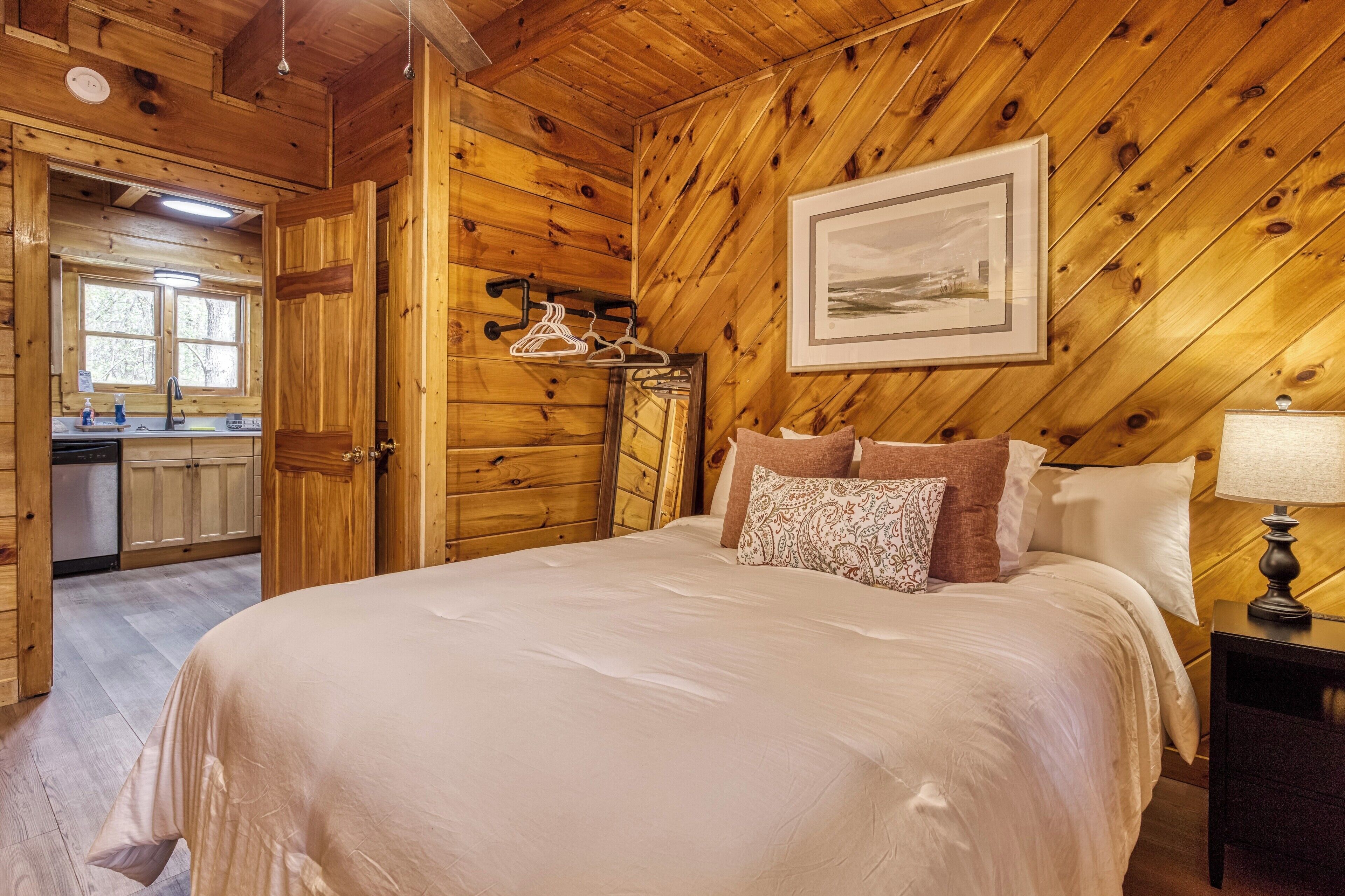 A wooden cabin interior with a bed, a nightstand, a framed picture, and a kitchen area.