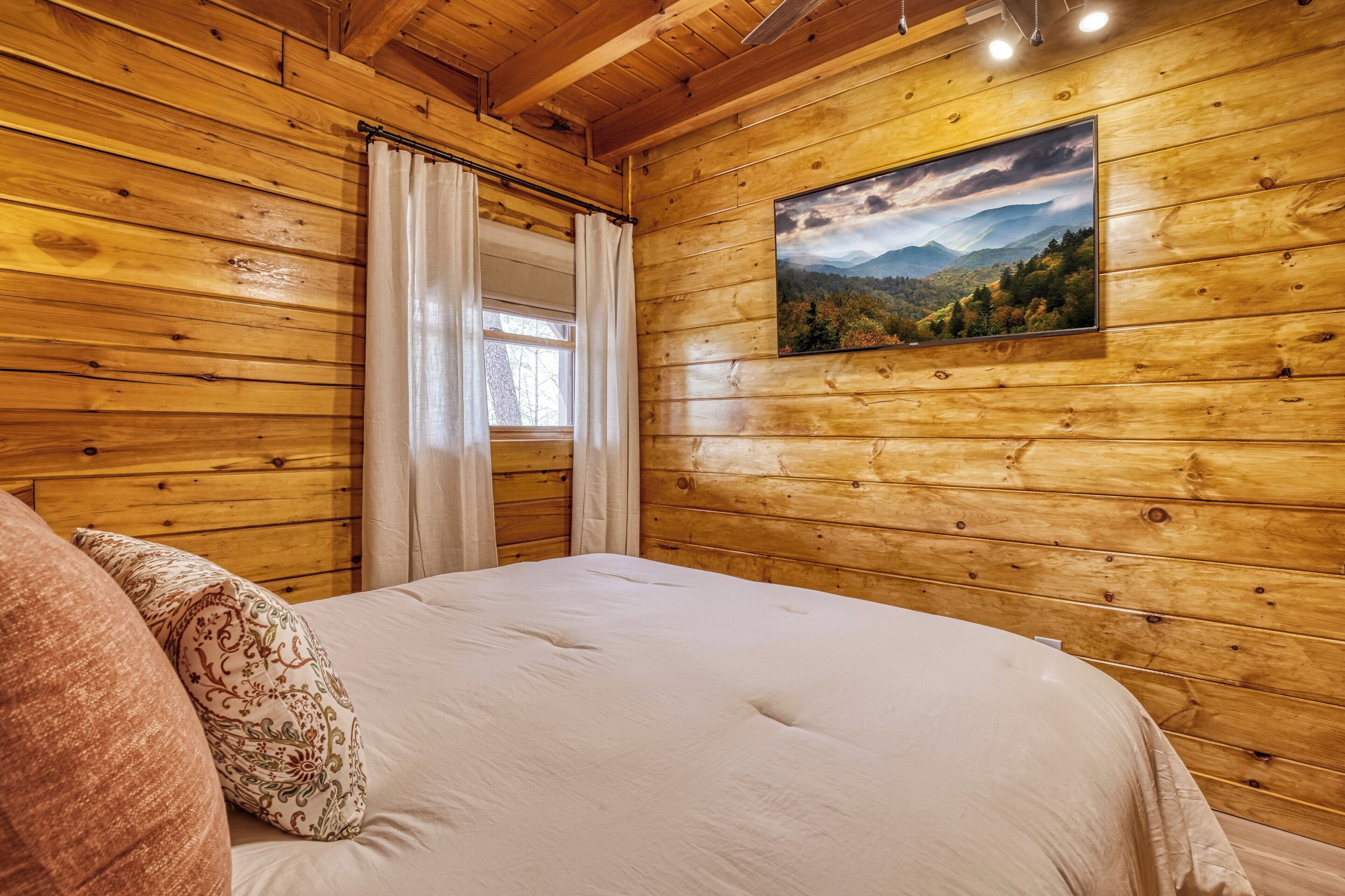 A wooden-paneled room with a bed, a window with curtains, and a landscape painting on the wall.
