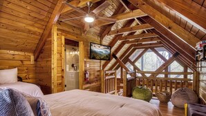 Interior de una cabaña de madera con una cama, un ventilador de techo, un cuadro y una ventana con vistas a árboles.