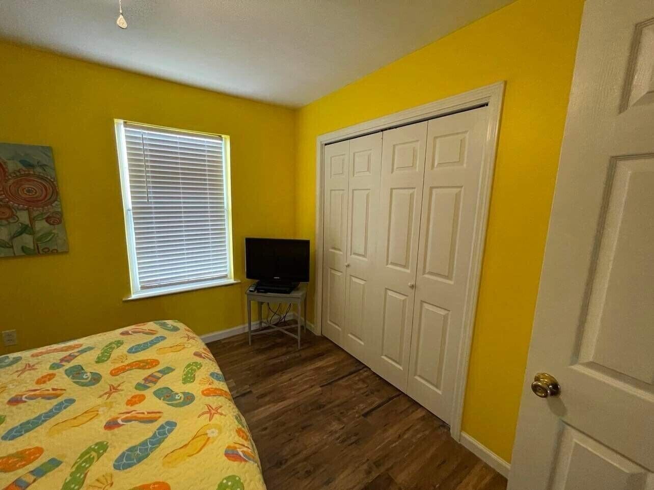 A bedroom with yellow walls, a bed with a colorful bedspread, a television on a small table, and a white closet.