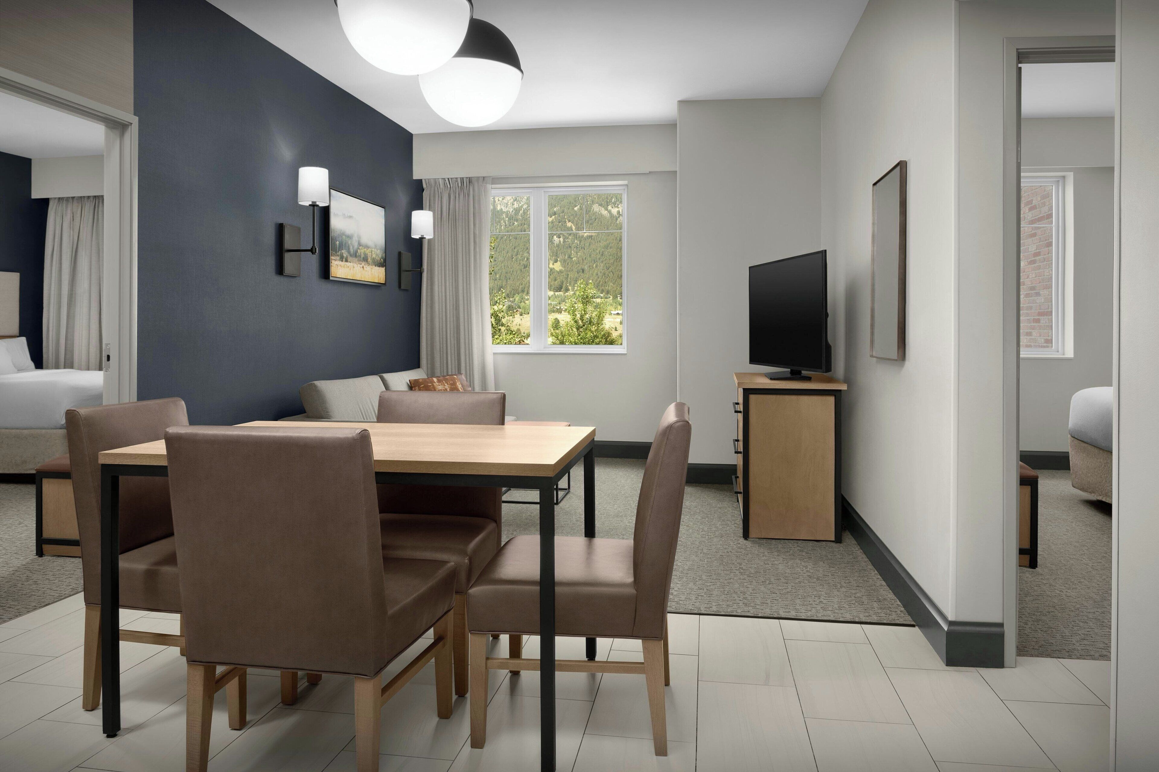 A modern hotel room with a dining area, a flat-screen TV, and a large window offering a view of greenery.