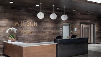 A reception desk at The Wilson Hotel with a wooden wall backdrop, a potted orchid, and three hanging crystal chandeliers.