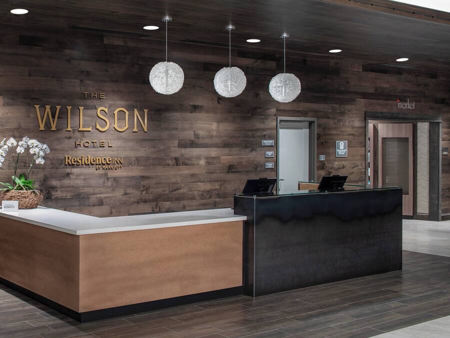 A reception desk at The Wilson Hotel with a wooden wall backdrop, a potted orchid, and three hanging crystal chandeliers.