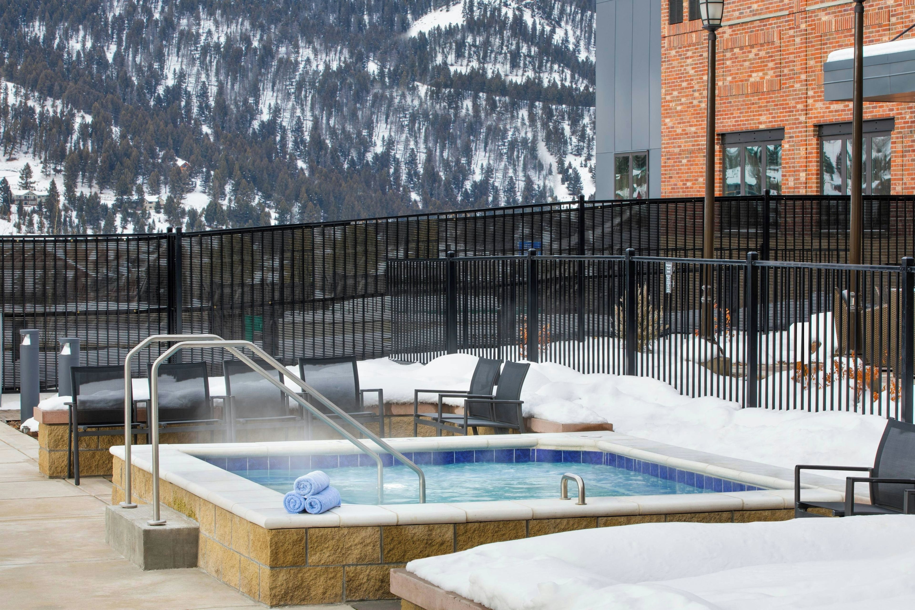 Ein beheizter Außenpool mit Stühlen und einer Sauna, umgeben von verschneiter Landschaft und einem Ziegelgebäude.