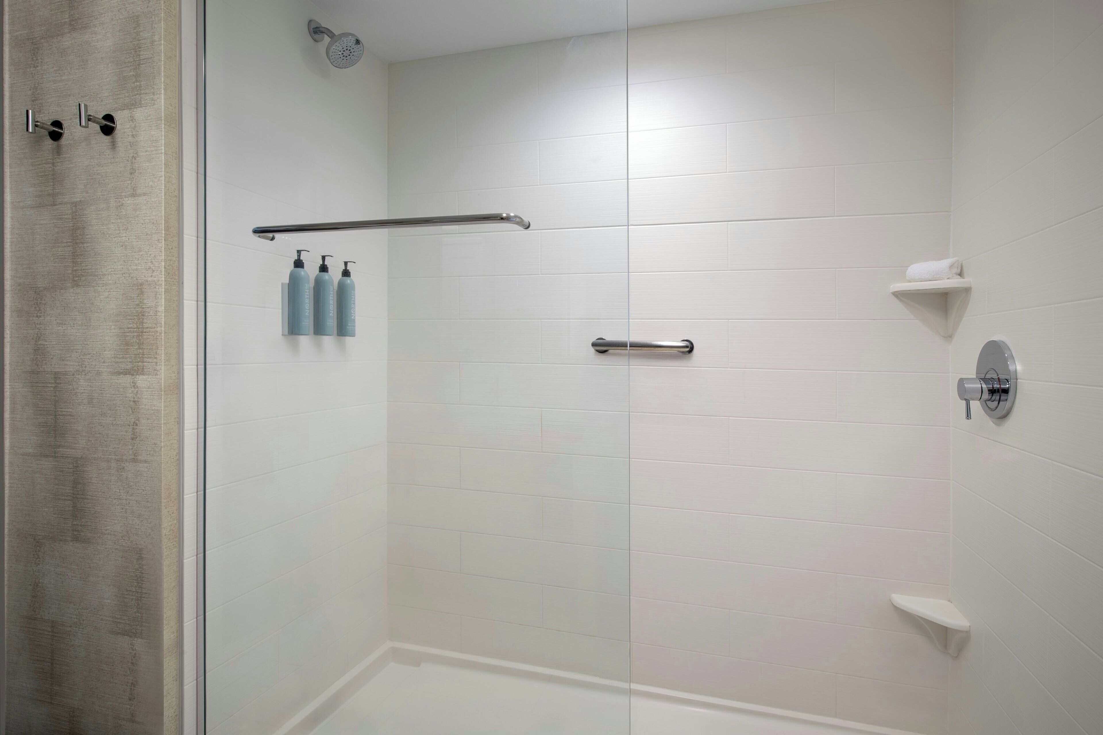A modern bathroom with white tiles, a glass shower enclosure, a towel rack, and a soap dispenser.