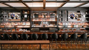 A bar area with multiple shelves of bottles, a long wooden table, and black chairs.