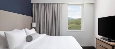 A hotel room with a bed, a television, a window with a view of a forest and mountains, and a dresser.