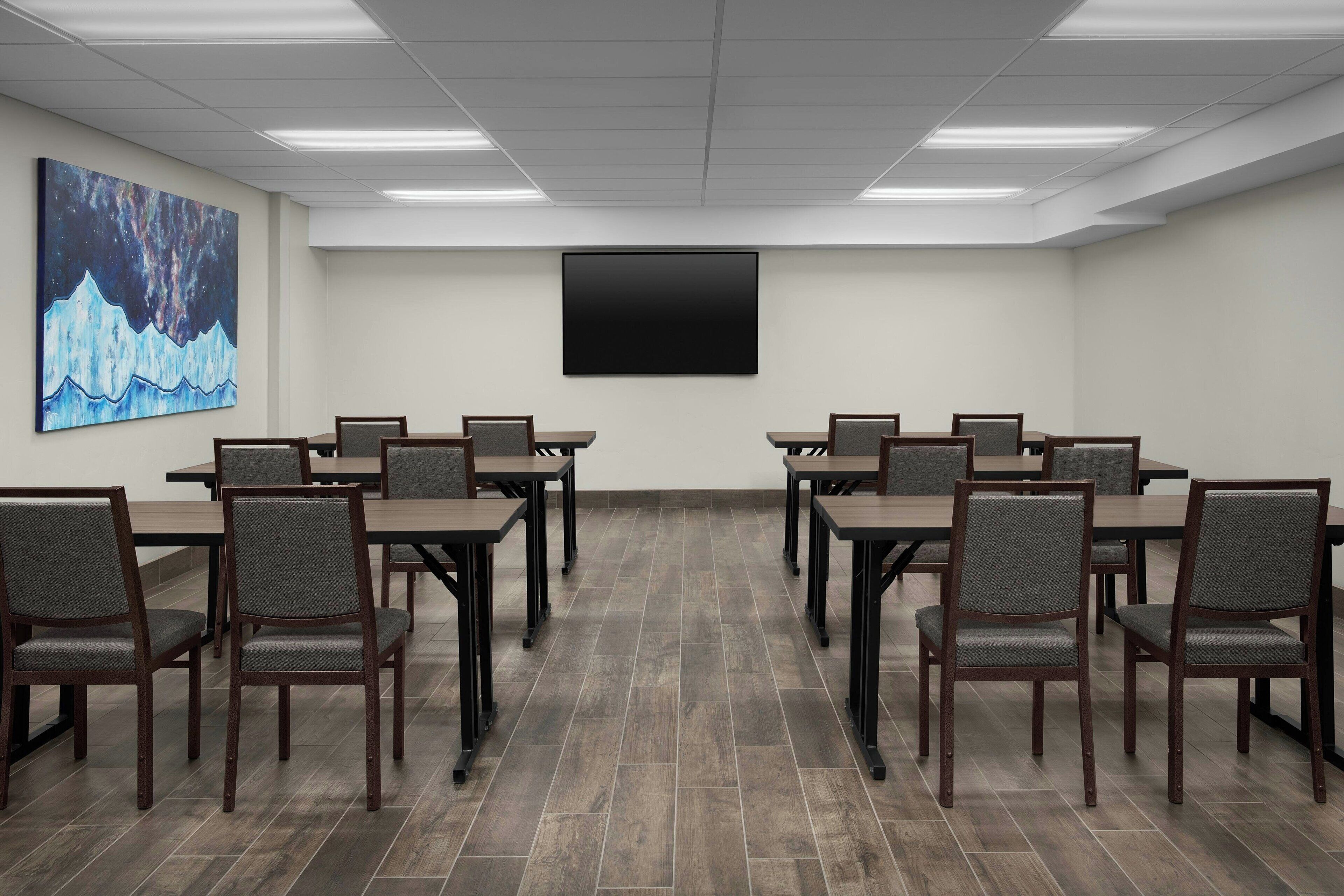 Un salón de conferencias con una amplia televisión de pantalla plana, mesas de madera y sillas tapizadas en gris.