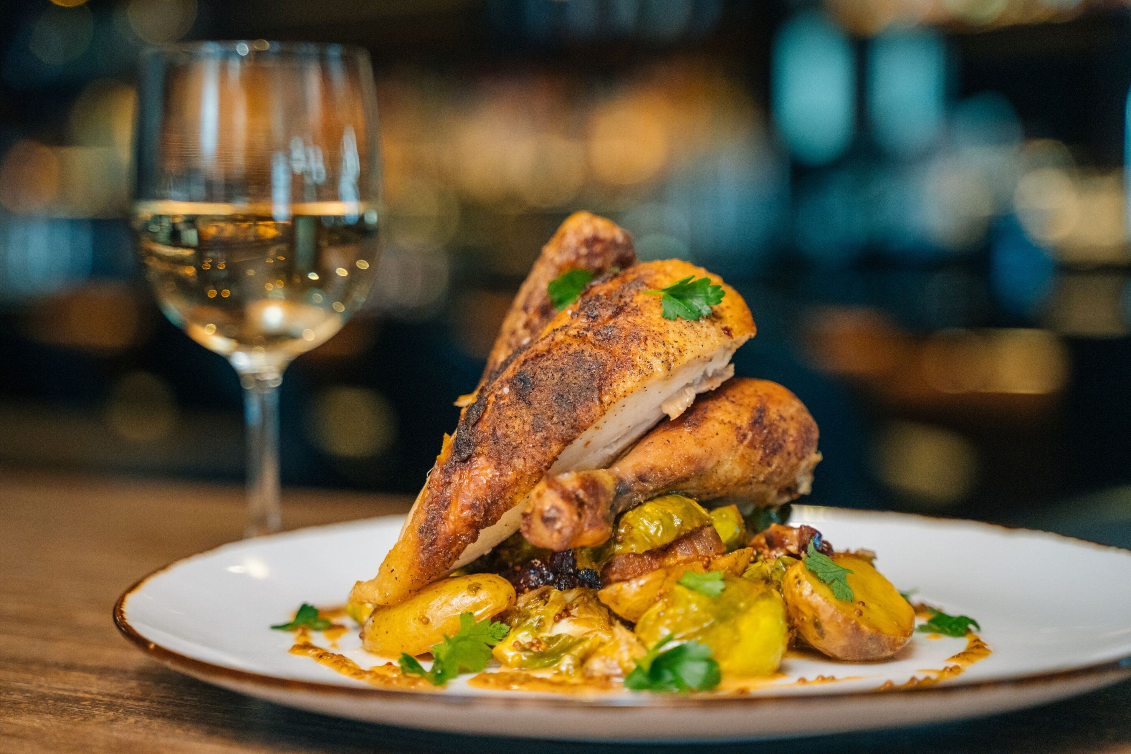 A plate of roasted chicken with vegetables and a glass of white wine on a wooden table.