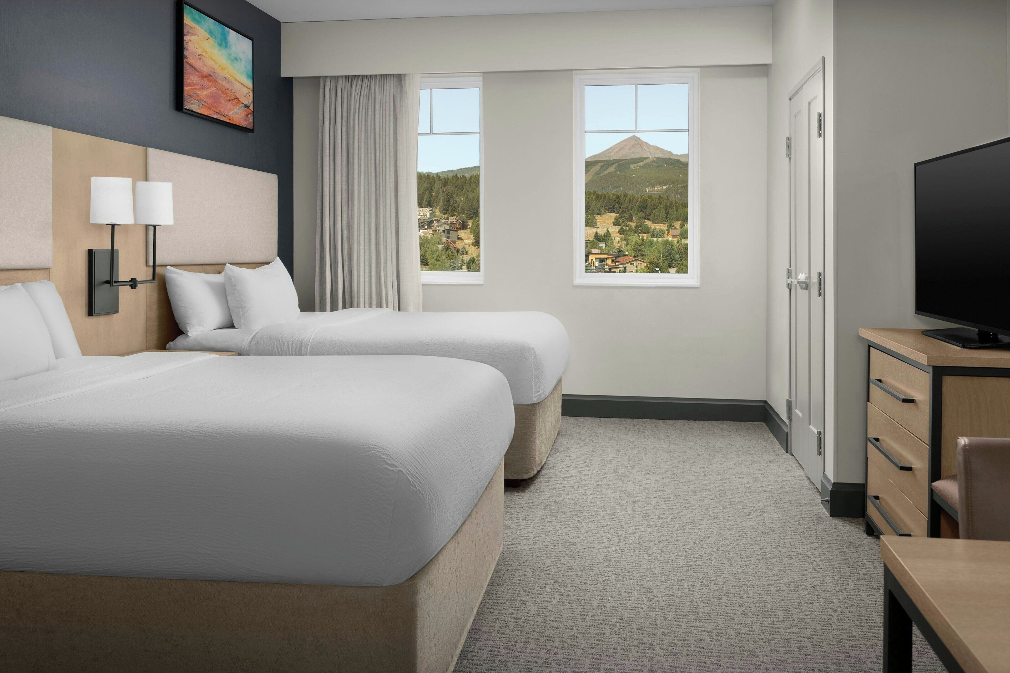 A hotel room with two beds, a TV, a desk, and a view of a mountain.