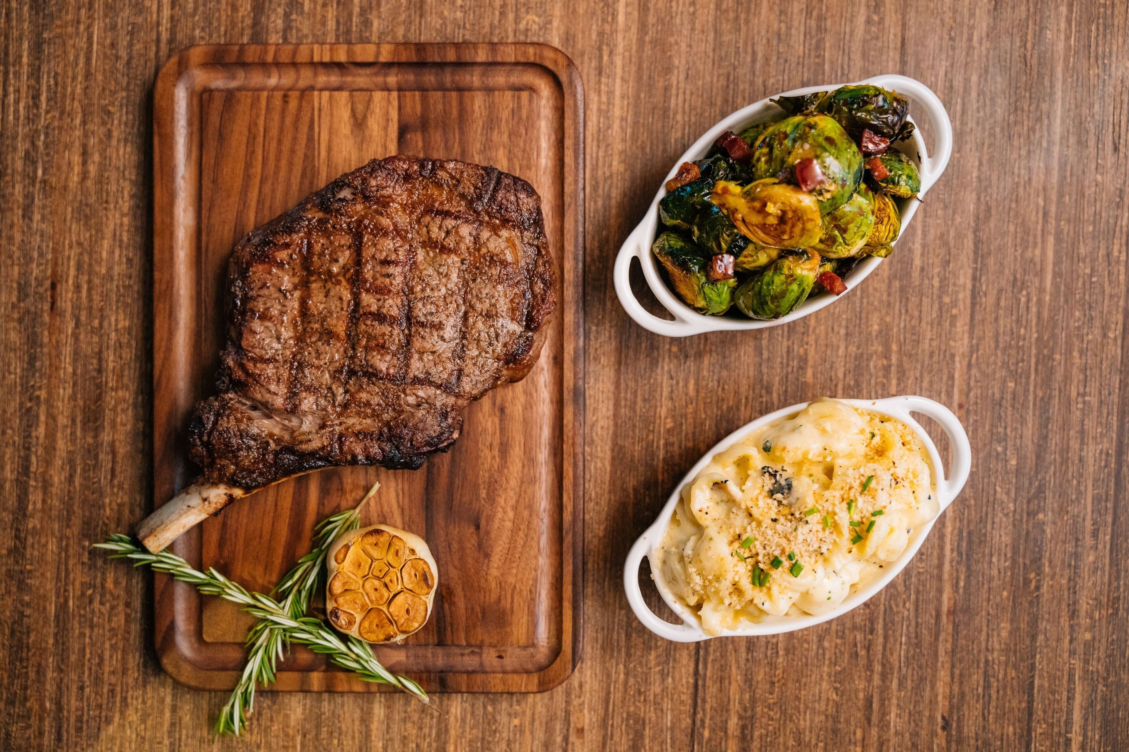 A steak on a wooden board, a dish of roasted Brussels sprouts, and a baked potato.