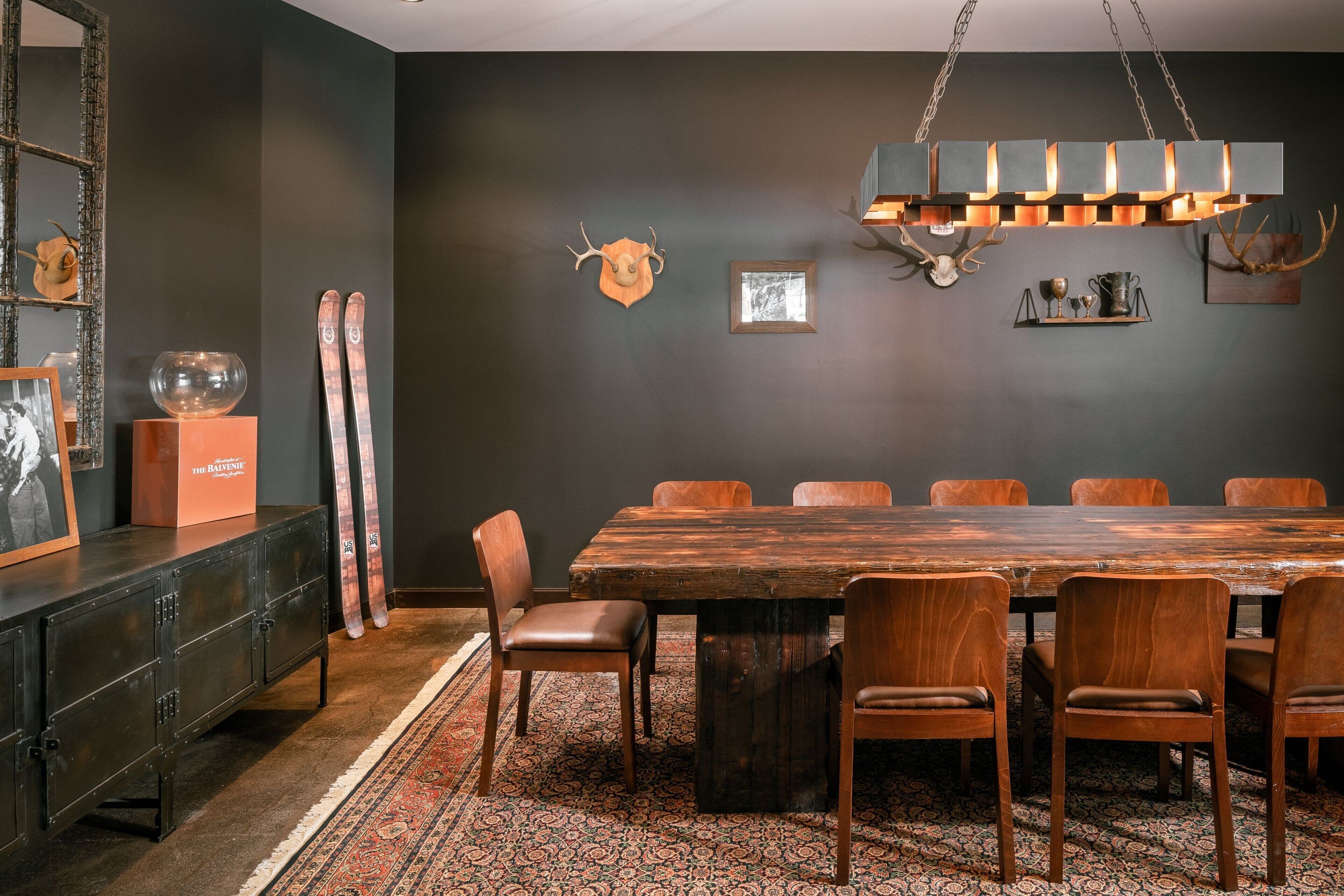Un coin repas avec une grande table en bois, six chaises, un lustre et des décorations murales, notamment une tête de cerf et des tableaux encadrés.