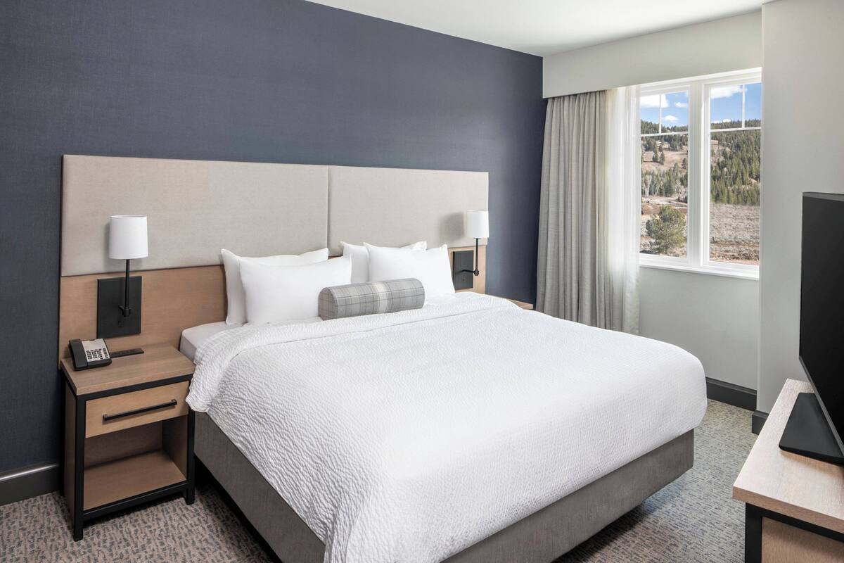 A hotel room with a large bed, two bedside tables with lamps, a flat-screen TV, and a window with a view of the outdoors.