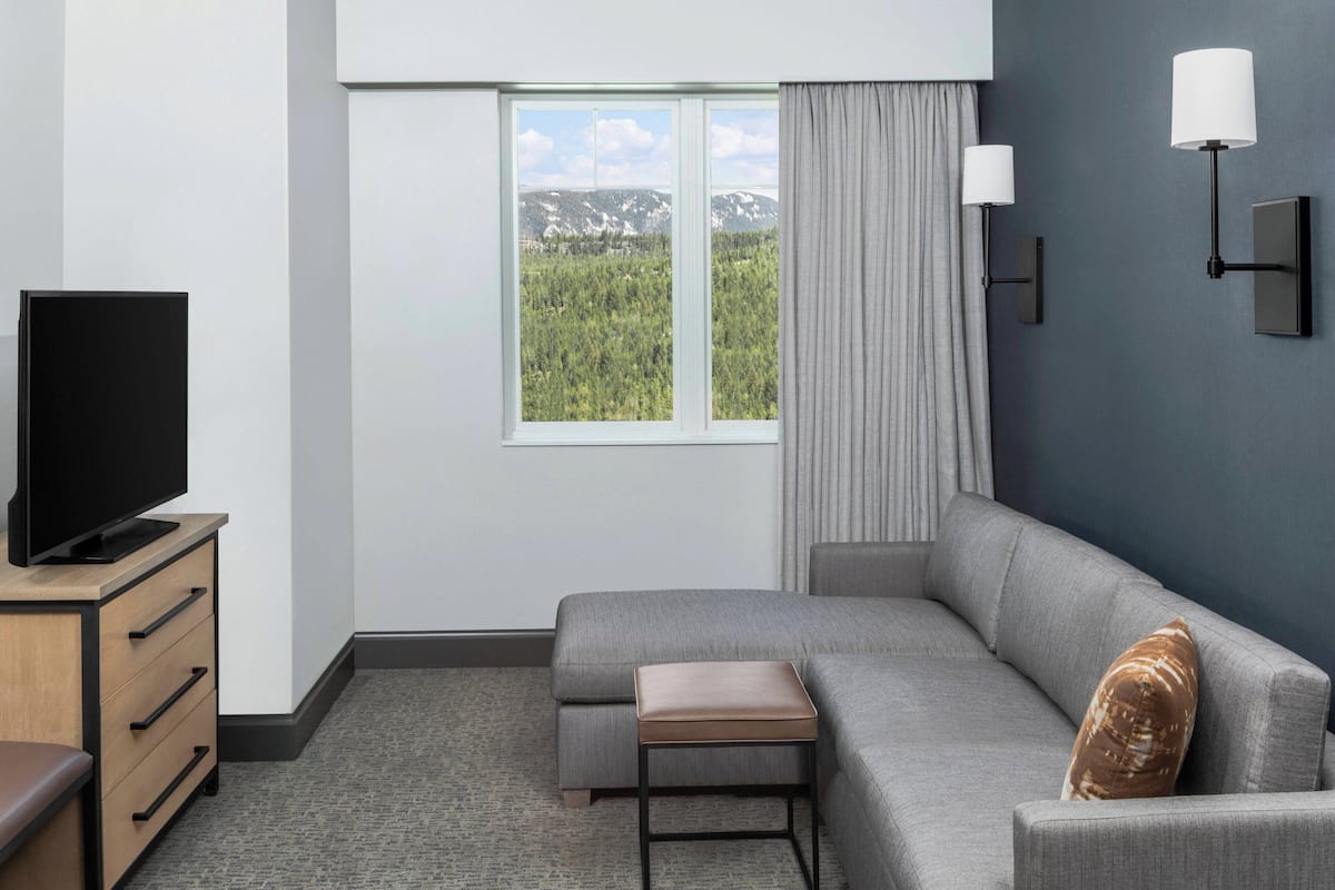 A modern hotel room with a large window, a flat-screen TV, a grey sofa, a small table, and a wall-mounted lamp.