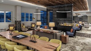A modern hotel lobby with a wooden reception desk, yellow chairs, a fireplace, and large windows offering views of the outdoors.