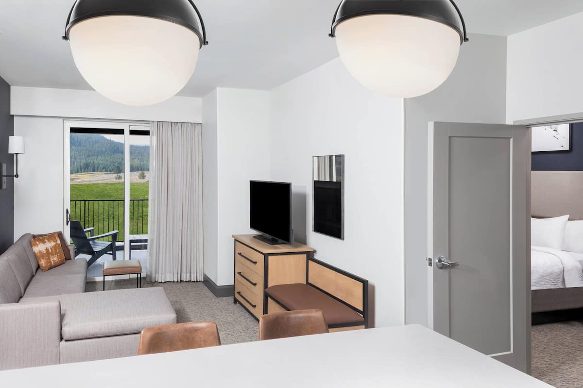 A modern hotel room with a sofa, a dining table, a TV, and a balcony with a view of the outdoors.