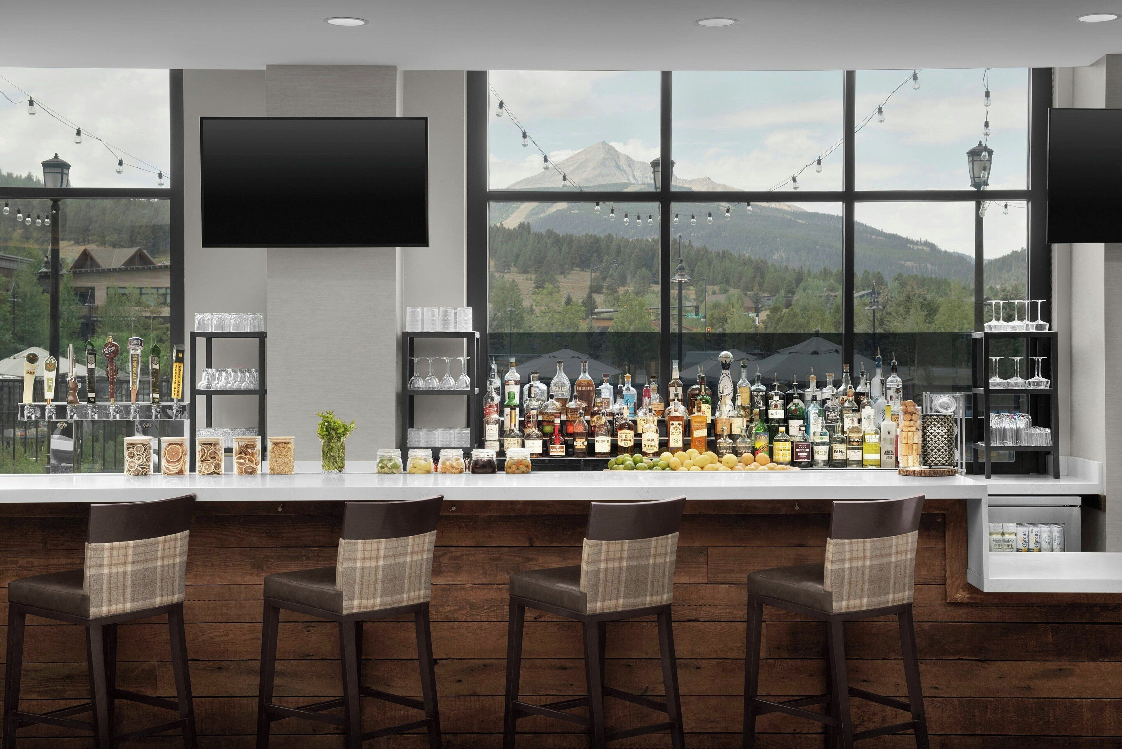 A bar area with a large window offering a mountain view, a flat-screen TV mounted above the bar, and a variety of bottles and glasses on the bar counter.
