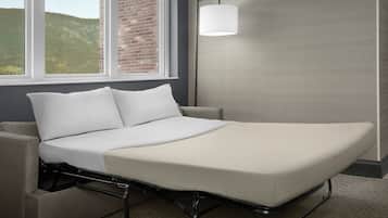 Una cama individual con estructura metálica, ropa de cama blanca y vistas a un muro de ladrillo y colinas a través de una ventana.