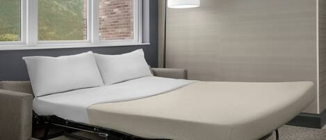 A single bed with a metal frame, white bedding, and a view of a brick wall and hills through a window.