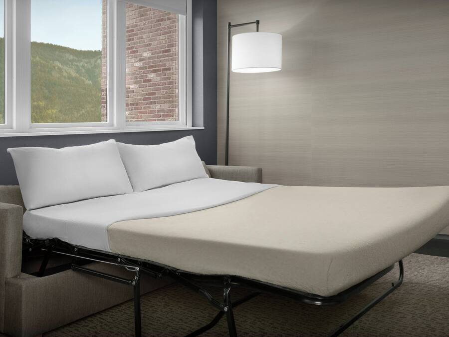 Una cama individual con estructura metálica, ropa de cama blanca y vistas a un muro de ladrillo y colinas a través de una ventana.